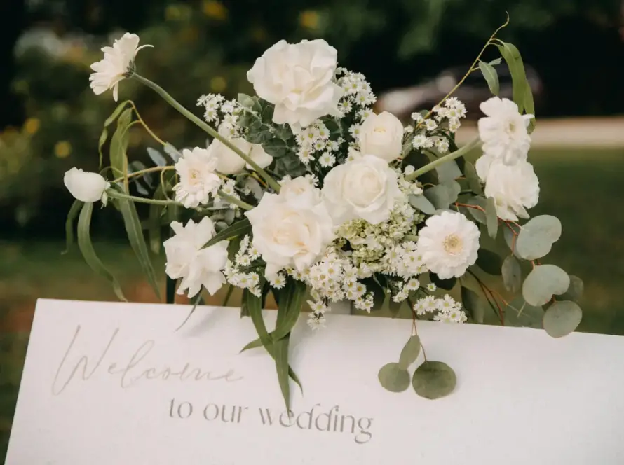 Elegant white floral arrangement above a wedding welcome sign reading “Welcome to our wedding,” set in an outdoor garden setting