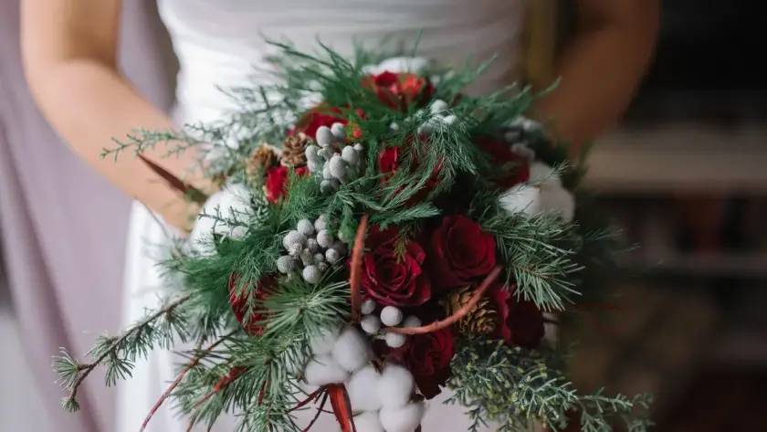 Bride holding a festive winter wedding bouquet featuring deep red roses, evergreen branches, pinecones, and silver berries, perfect for a holiday wedding in Orlando.