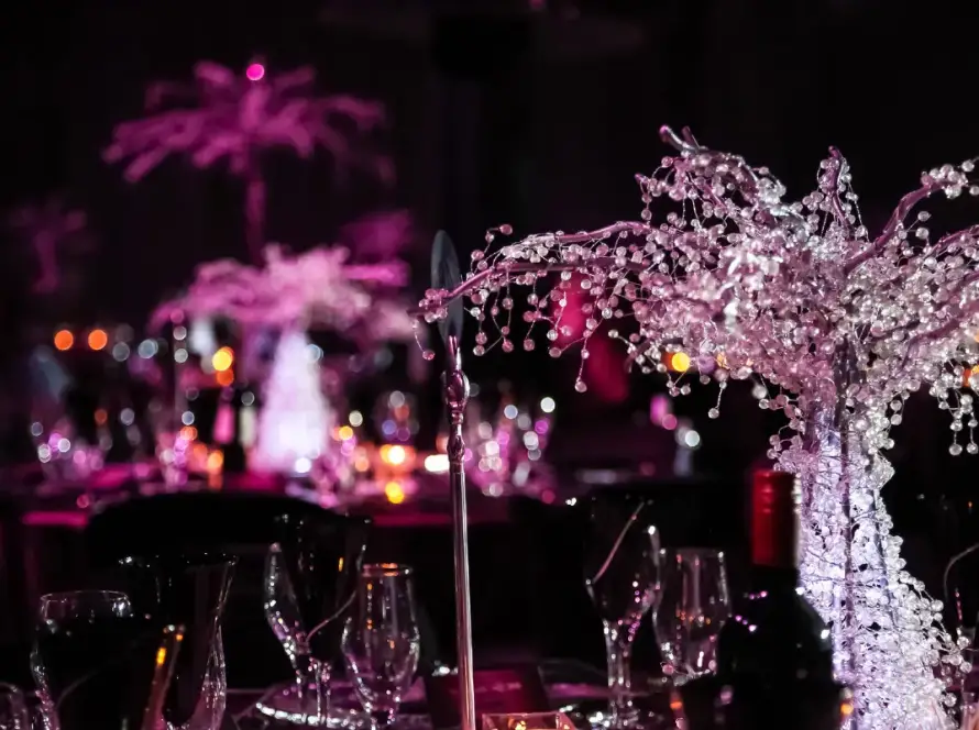 Elegant gala table setting with crystal-like decorative centerpieces, soft candlelight, and pink ambient lighting creating a luxurious event atmosphere