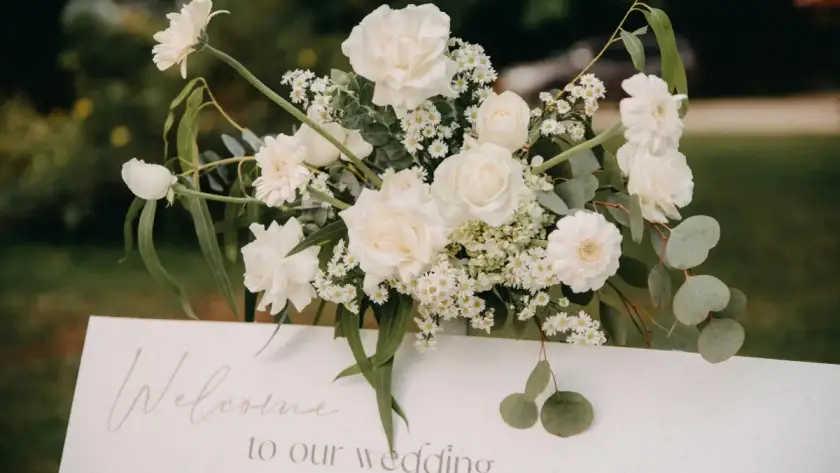 Elegant white floral arrangement above a wedding welcome sign reading “Welcome to our wedding,” set in an outdoor garden setting