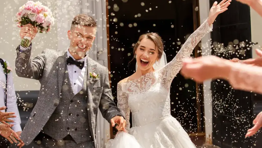 Joyful bride and groom celebrating outside their wedding ceremony, smiling and raising their arms as guests toss confetti around them