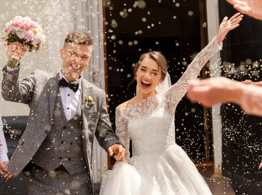 Joyful bride and groom celebrating outside their wedding ceremony, smiling and raising their arms as guests toss confetti around them