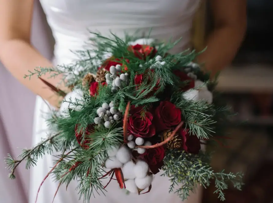 Bride holding a festive winter wedding bouquet featuring deep red roses, evergreen branches, pinecones, and silver berries, perfect for a holiday wedding in Orlando.